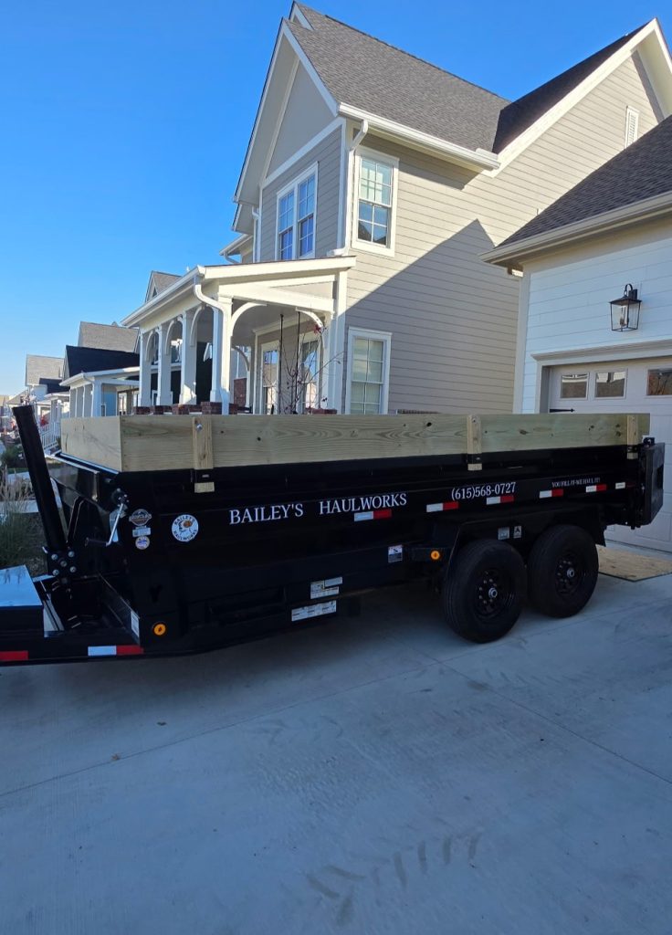 Black utility trailer labeled ‘Bailey’s Haulworks’ parked in a driveway in front of a suburban house.